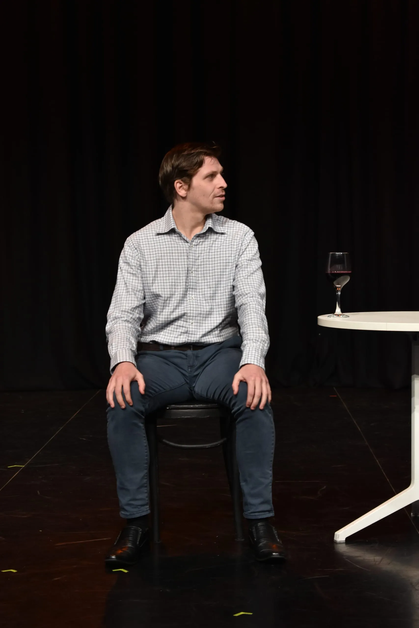A performer sits alone at a small white café table against a black curtain, a glass of red wine at hand, glancing off stage