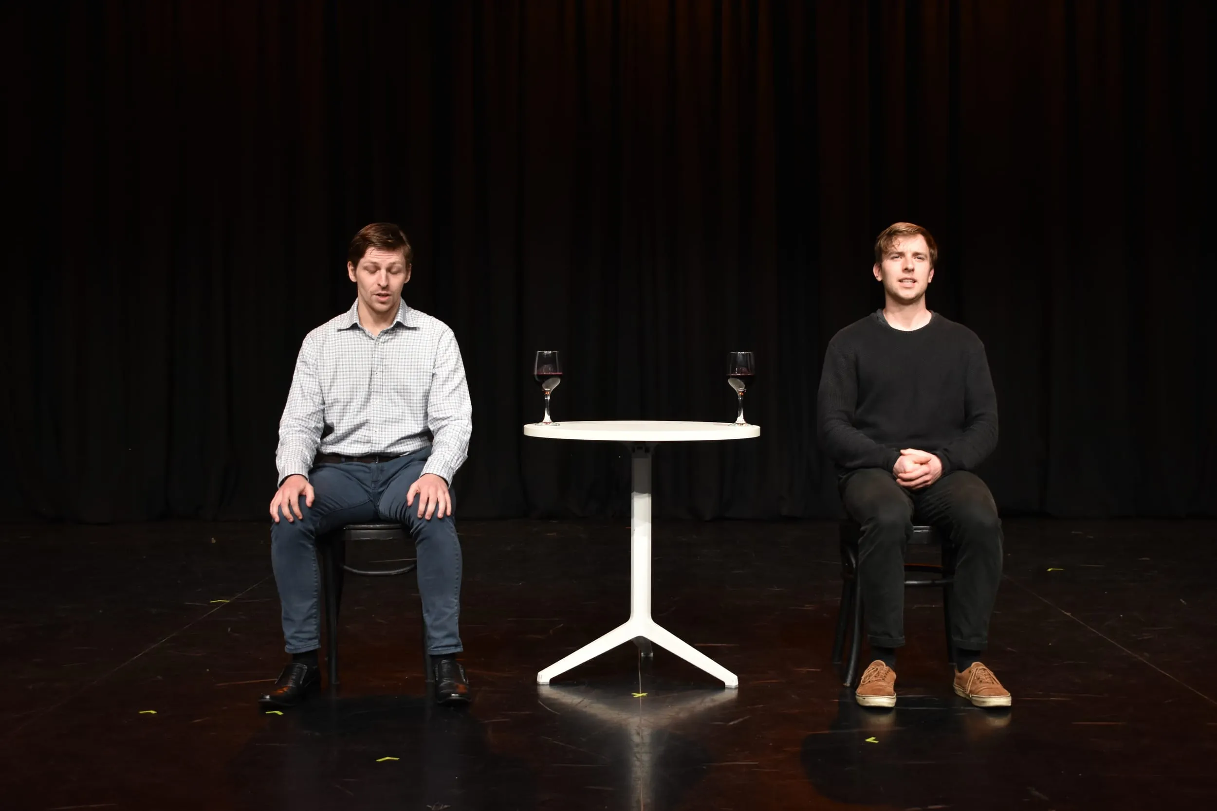 The two performers sit either side of the café table facing the audience, a glass of red wine at each place