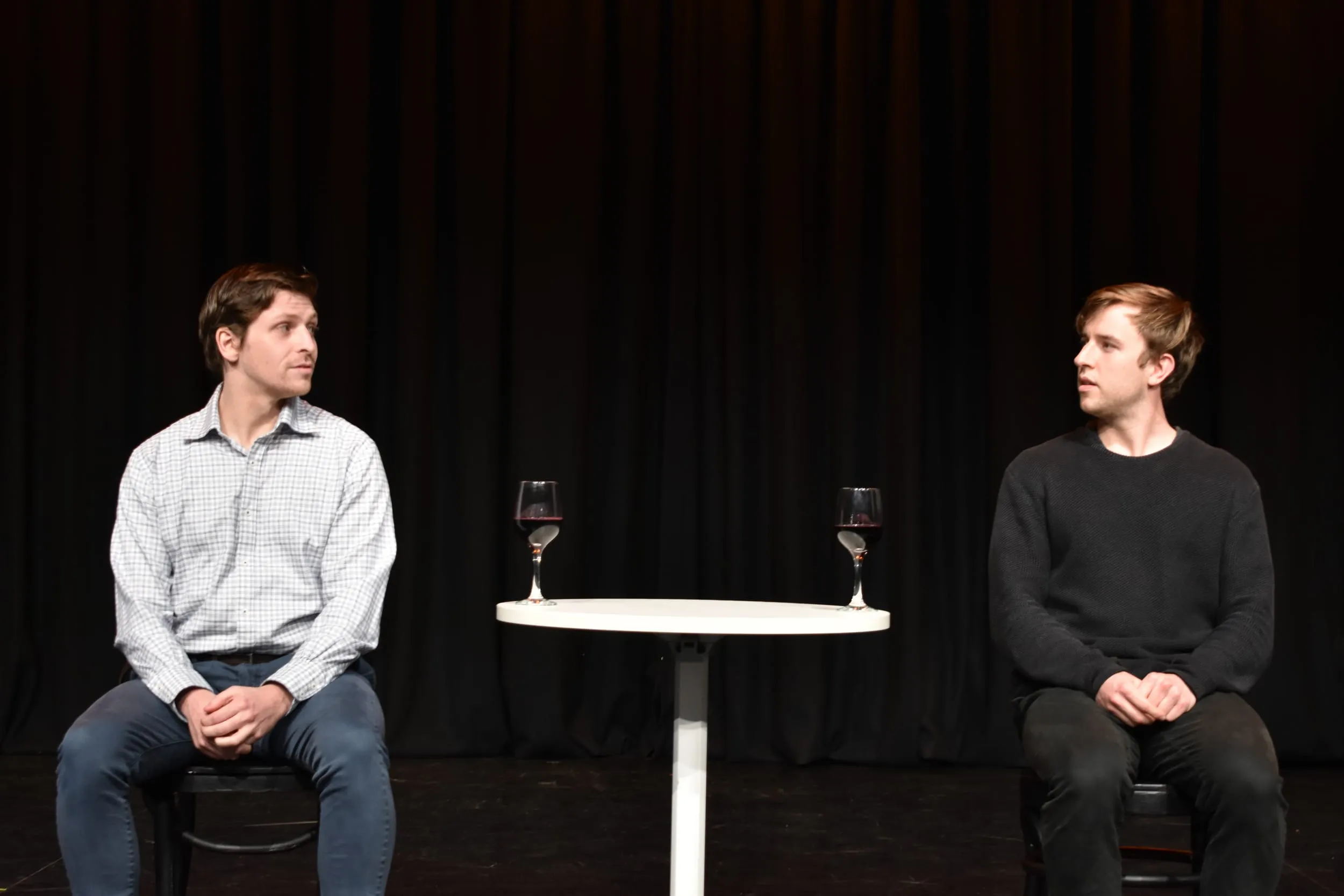 The two performers turn to face each other across the café table, mid-conversation