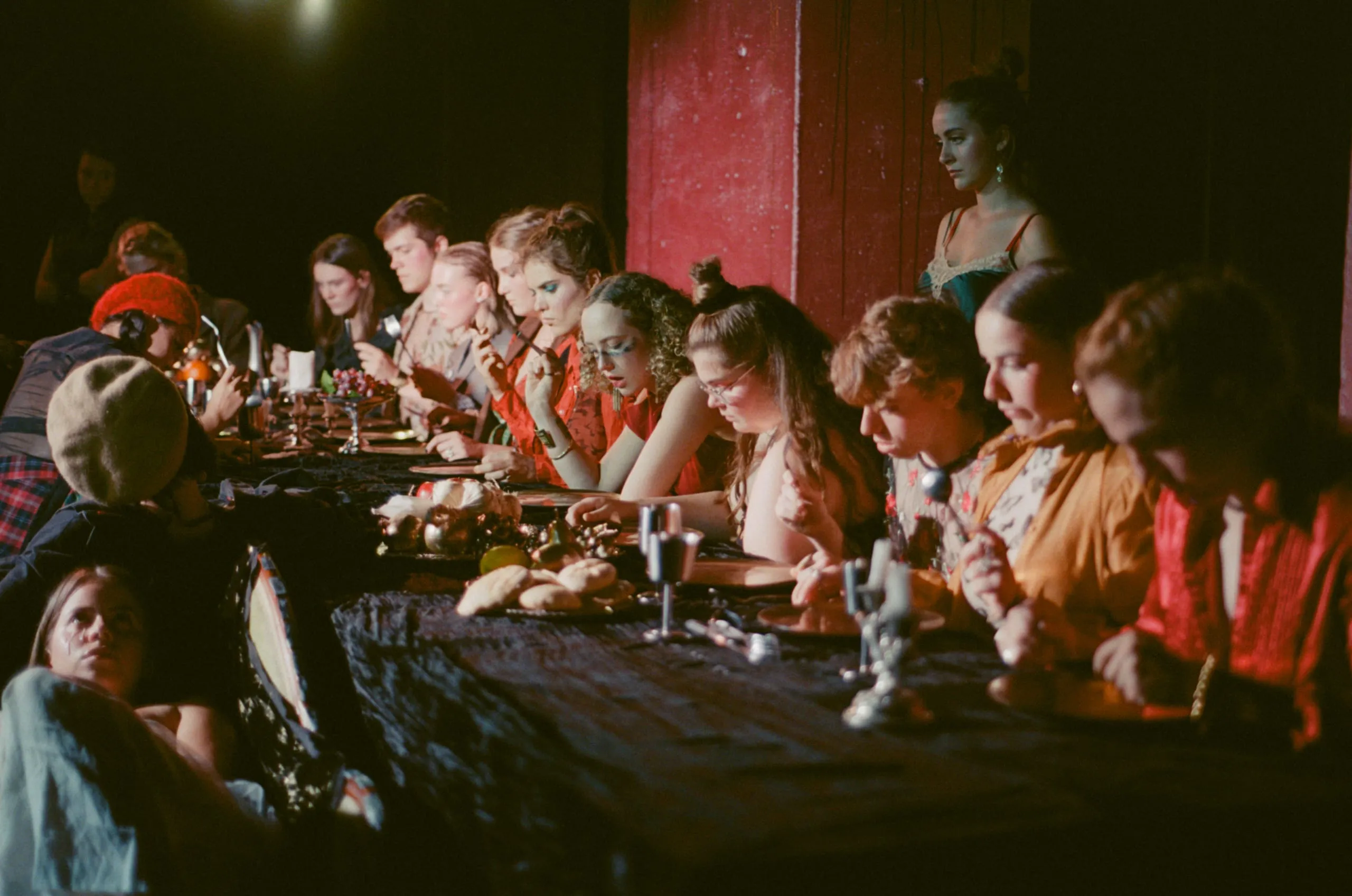 The ensemble picks at fruit and bread along a black-draped banqueting table against a red wall, one woman standing apart
