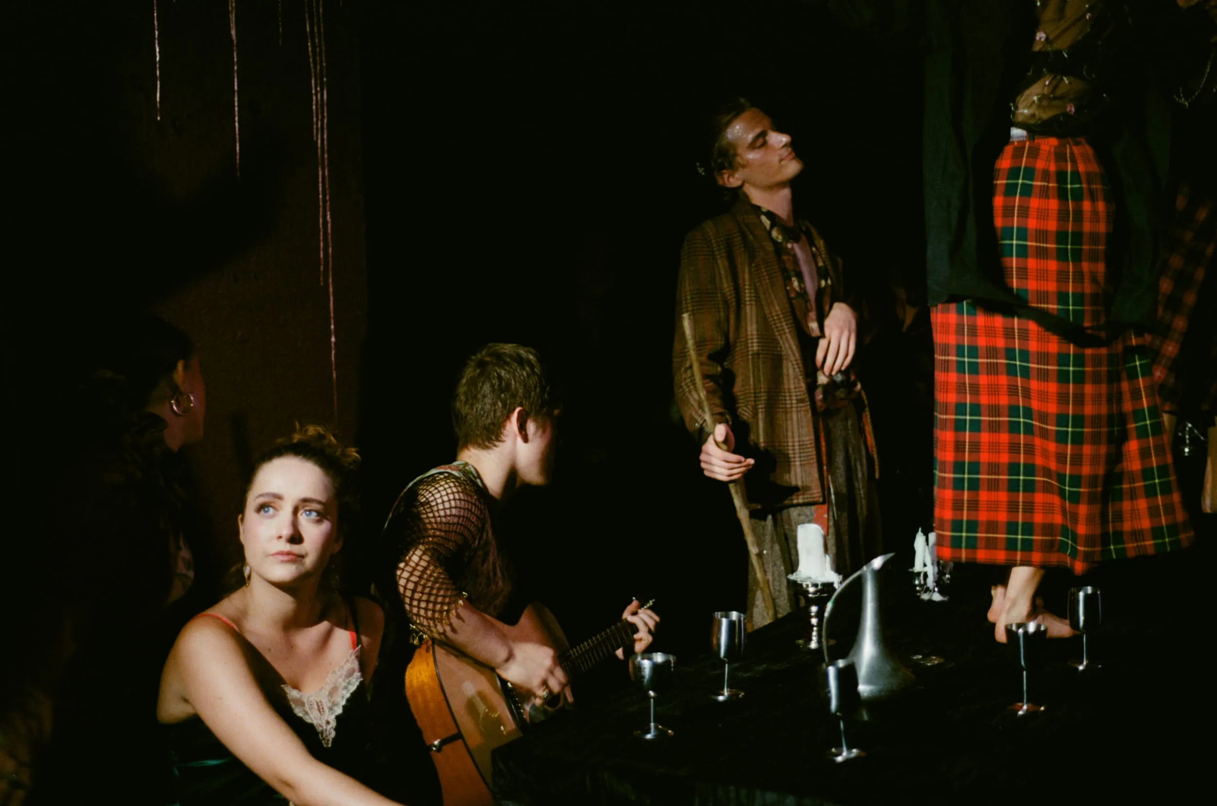 A candle-lit feast scene: a woman in a lace-trimmed slip gazes away while a guitarist plays and figures in tartan crowd the table