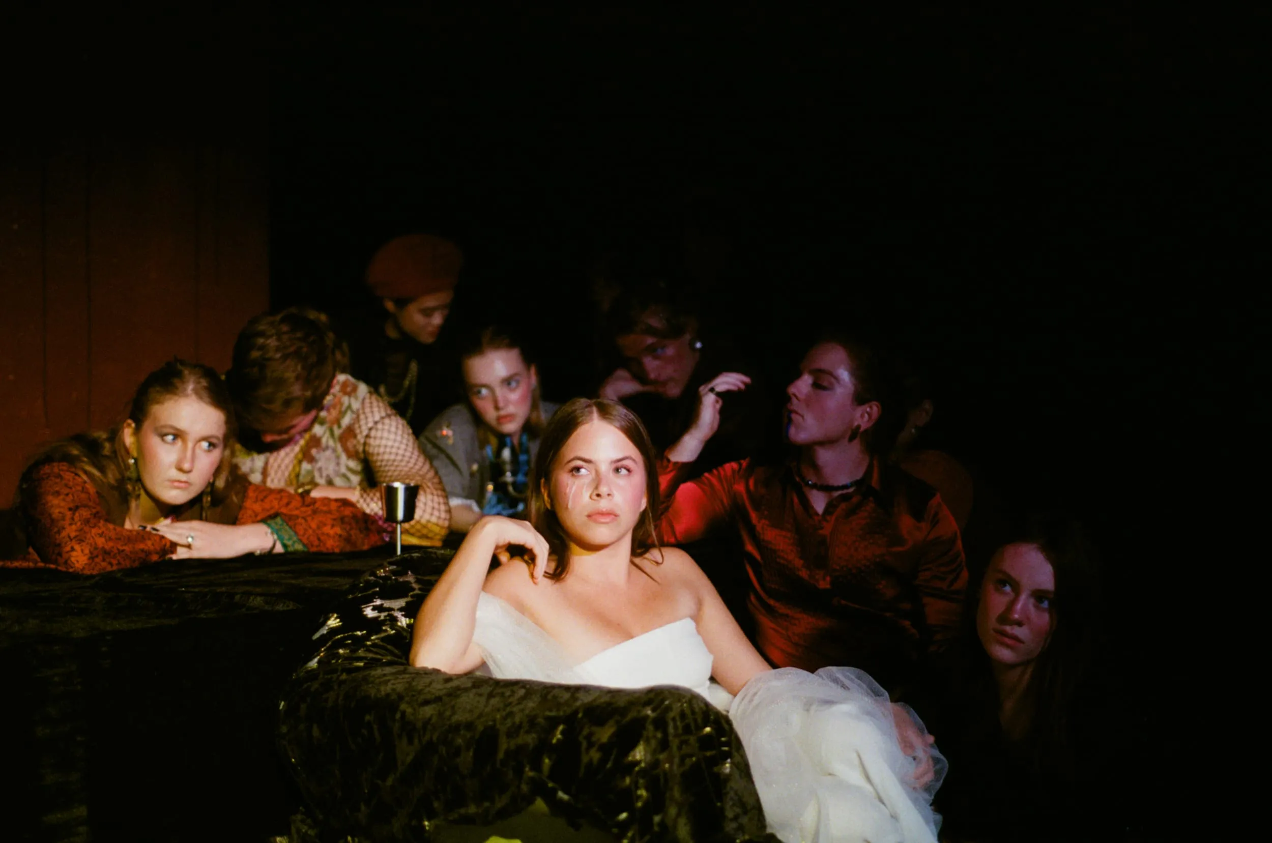 A woman in a white strapless gown stares out from a velvet-draped table as onlookers press in around her in the dark