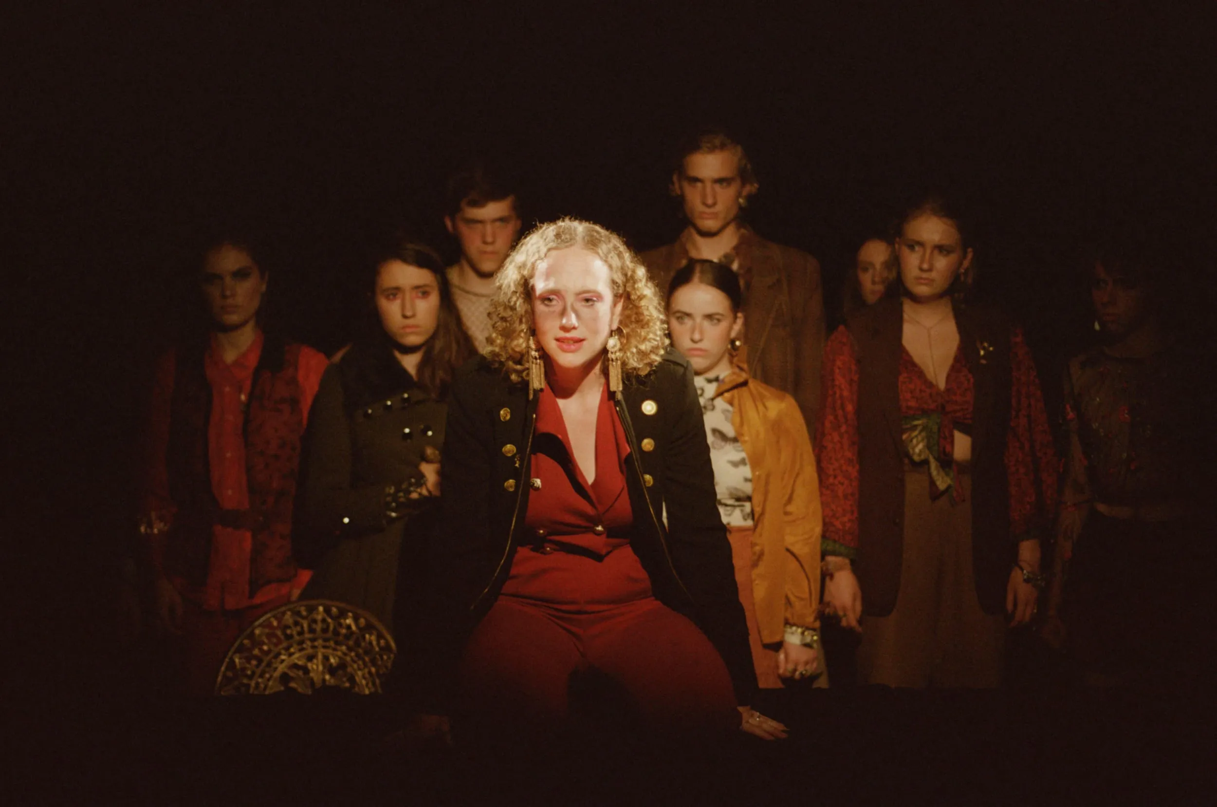 Caligula, curly-haired in a red suit and dark military coat, sits centre stage in warm light with the ensemble shadowed behind