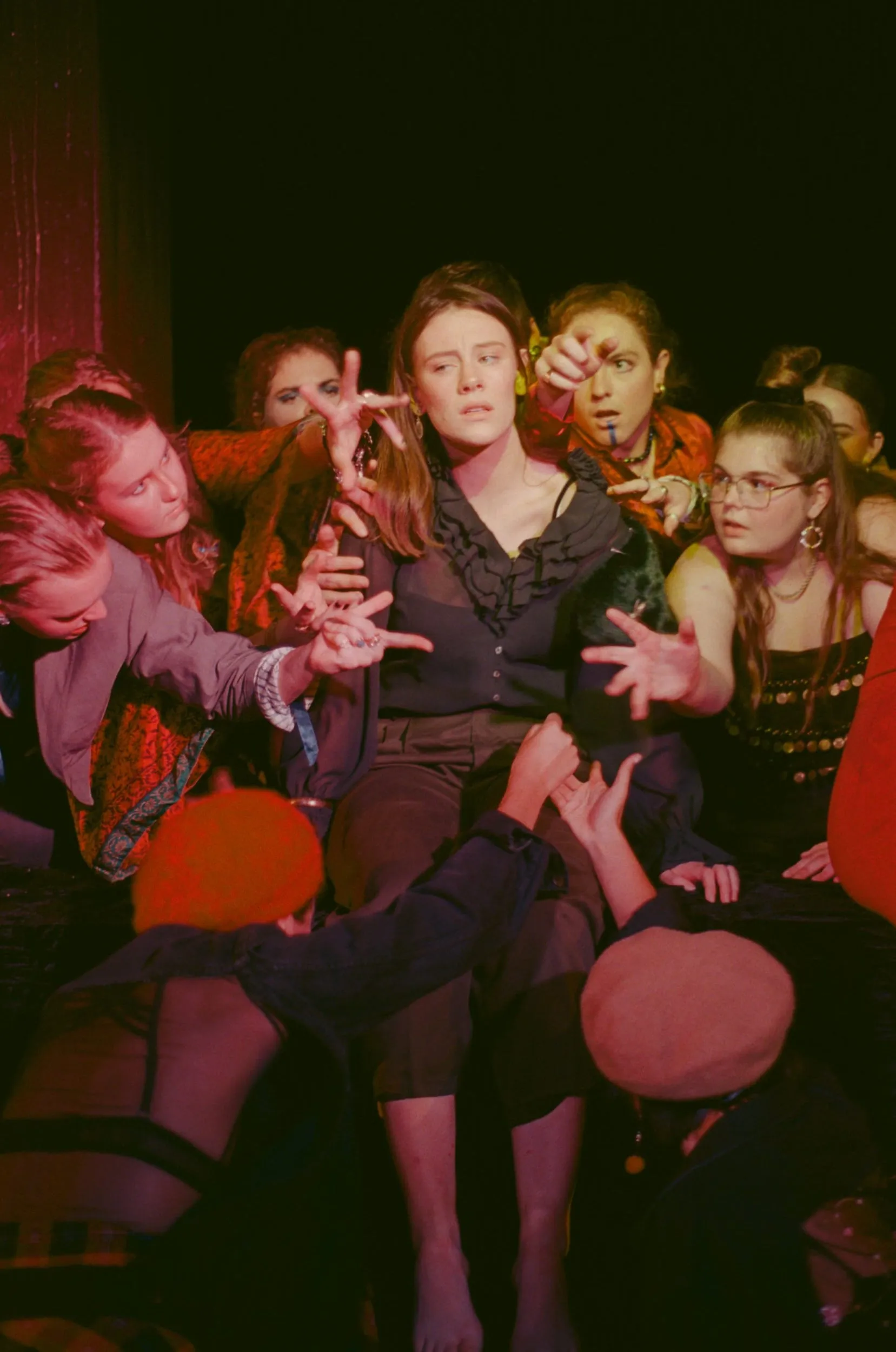 The ensemble converges on a seated woman, hands clawing towards her from every side under red light