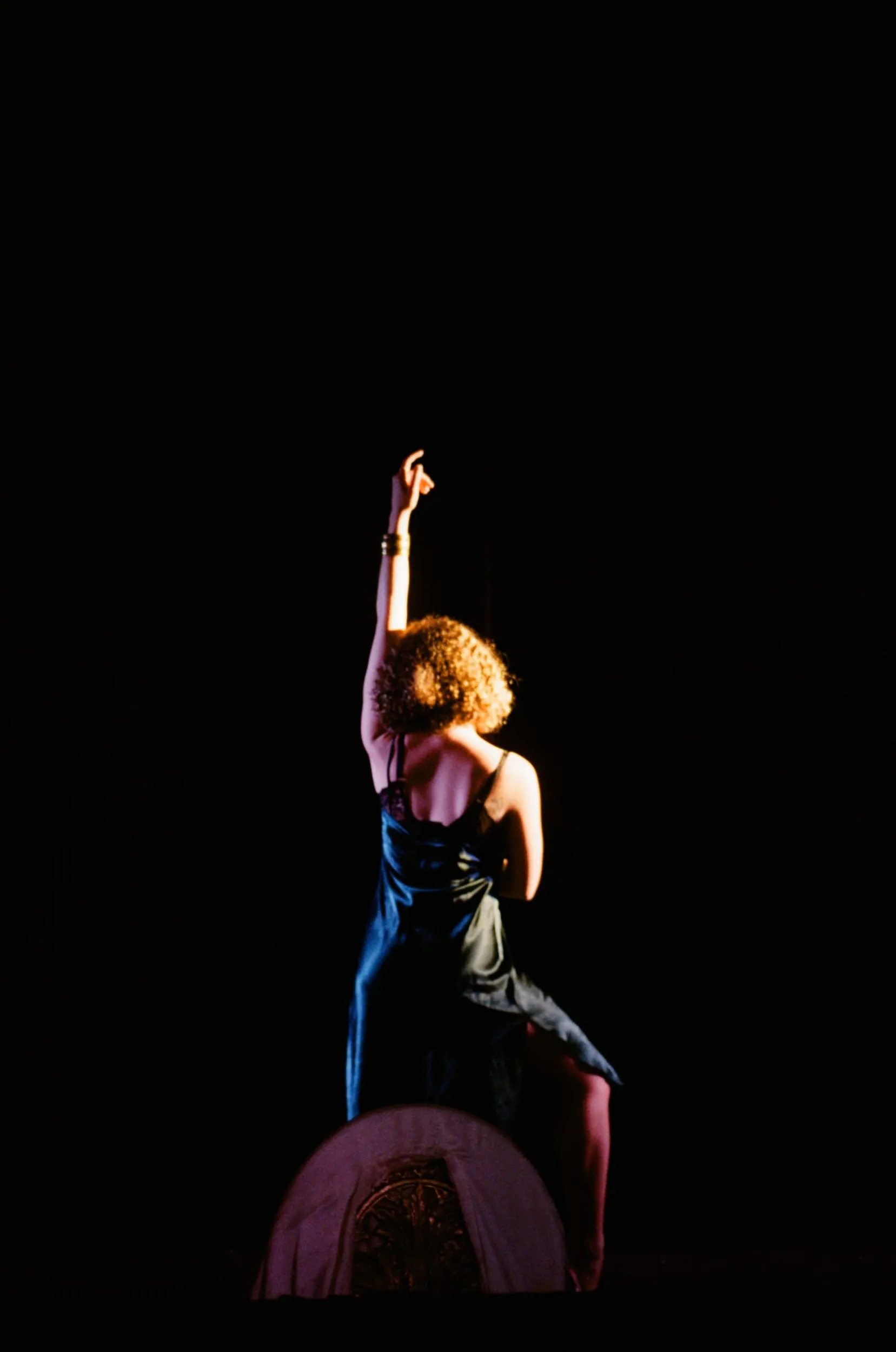 Seen from behind, a performer in a velvet slip reaches one arm to the sky in a pool of light