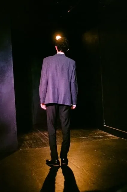 A suited figure stands with his back to the audience in a narrow shaft of warm light