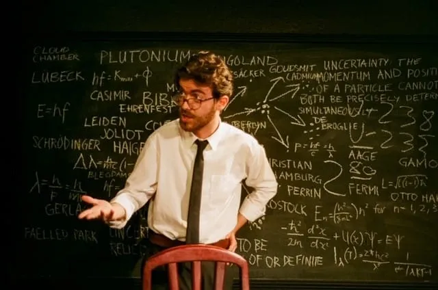 An actor gestures mid-argument before a blackboard scrawled with plutonium diagrams, formulas and physicists' names