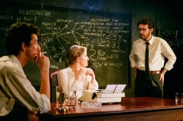 Margrethe sits at the typewriter between the two physicists as the debate continues across the blackboard