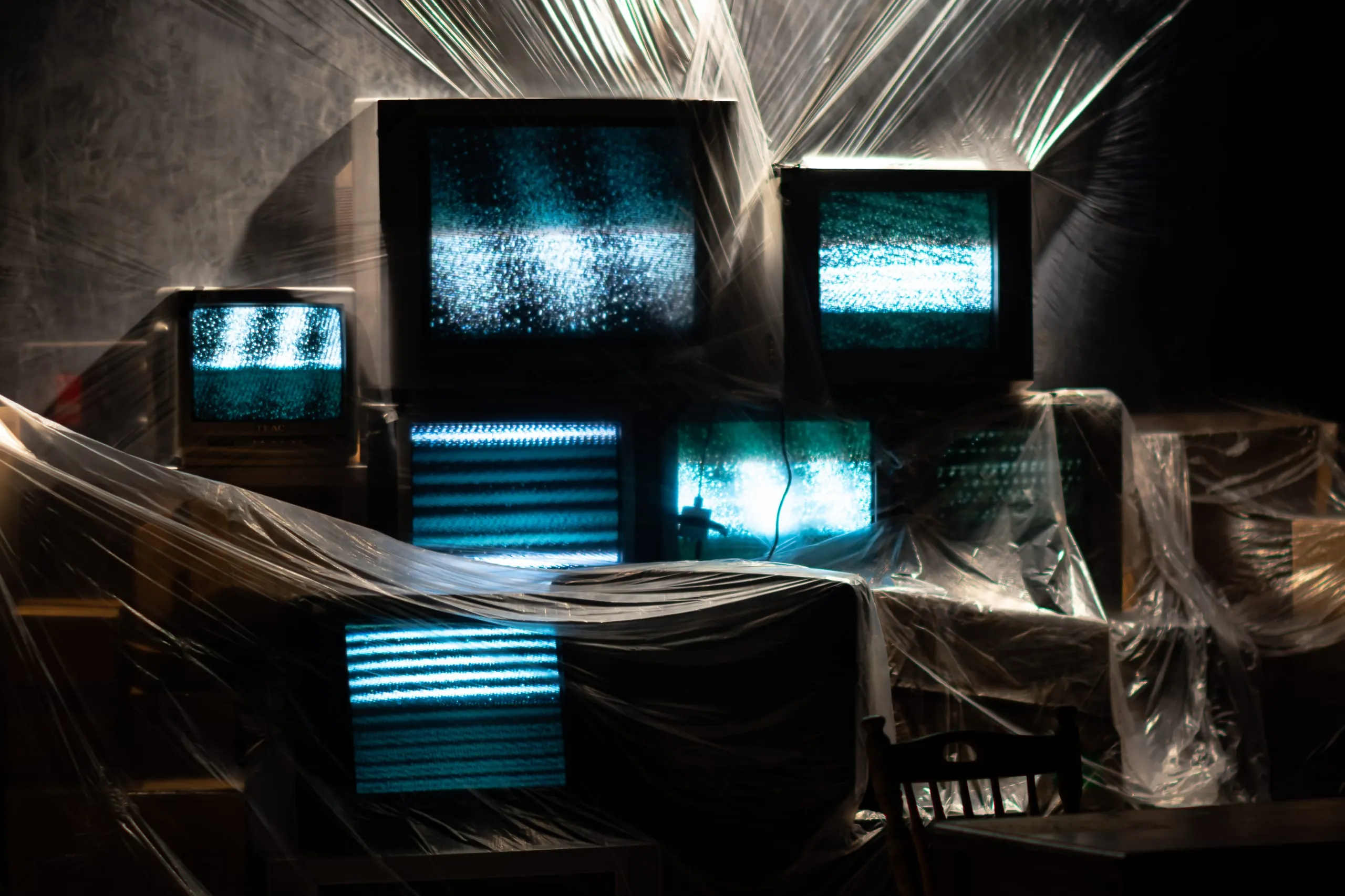 A bank of CRT televisions glows with blue static beneath sheets of plastic film