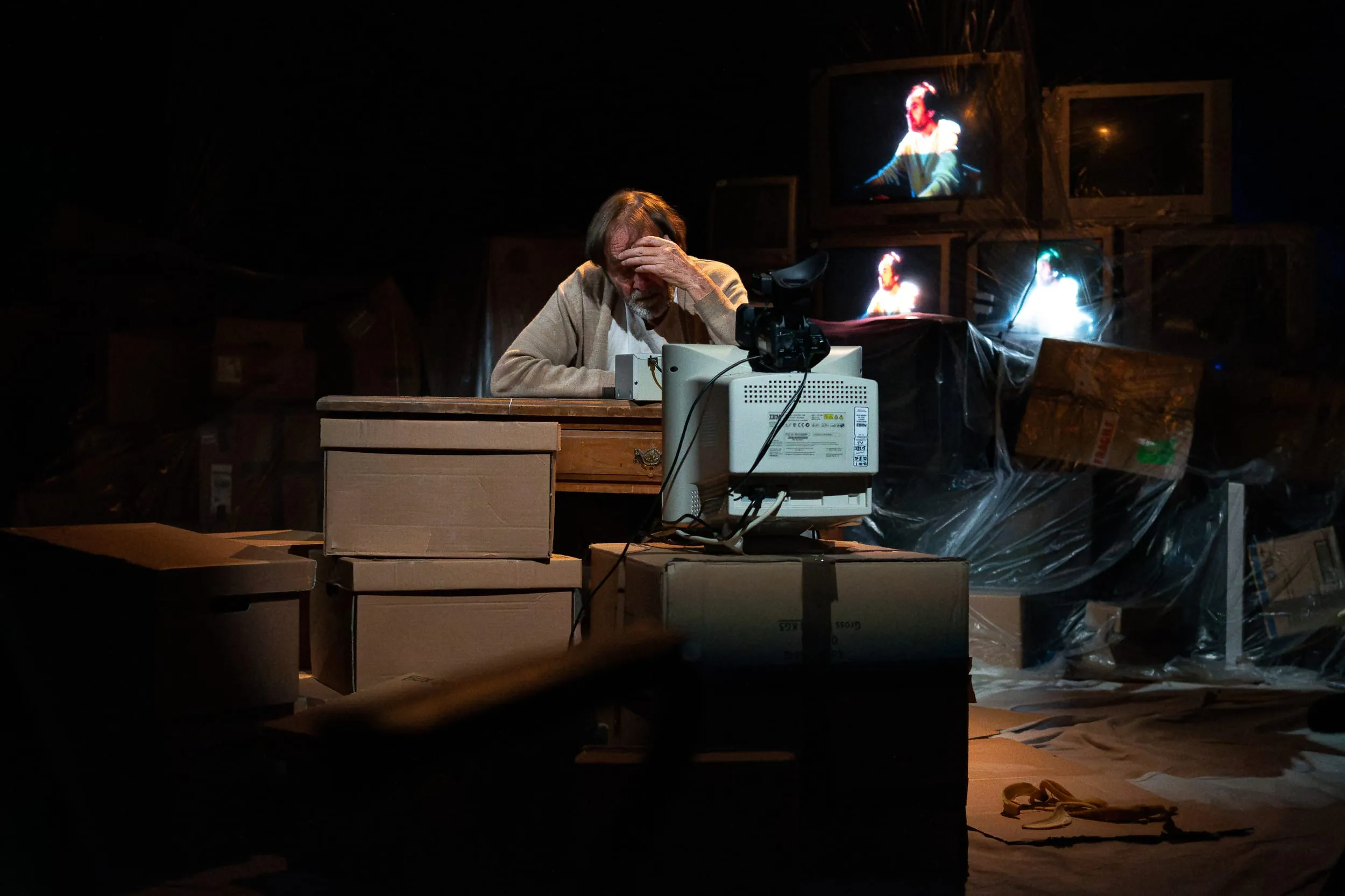 Krapp slumps behind an old CRT monitor with his head in his hand, hemmed in by cardboard boxes and shrouded televisions