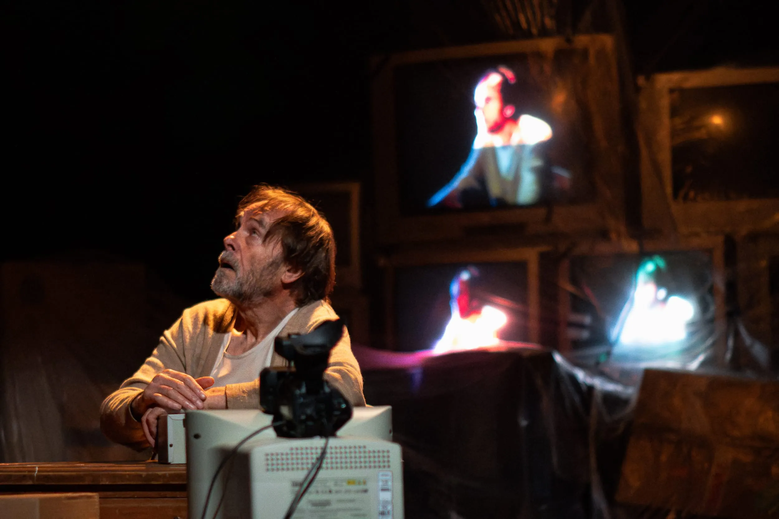 Krapp looks up past the camera mounted on his monitor as faces glow on the screens behind him