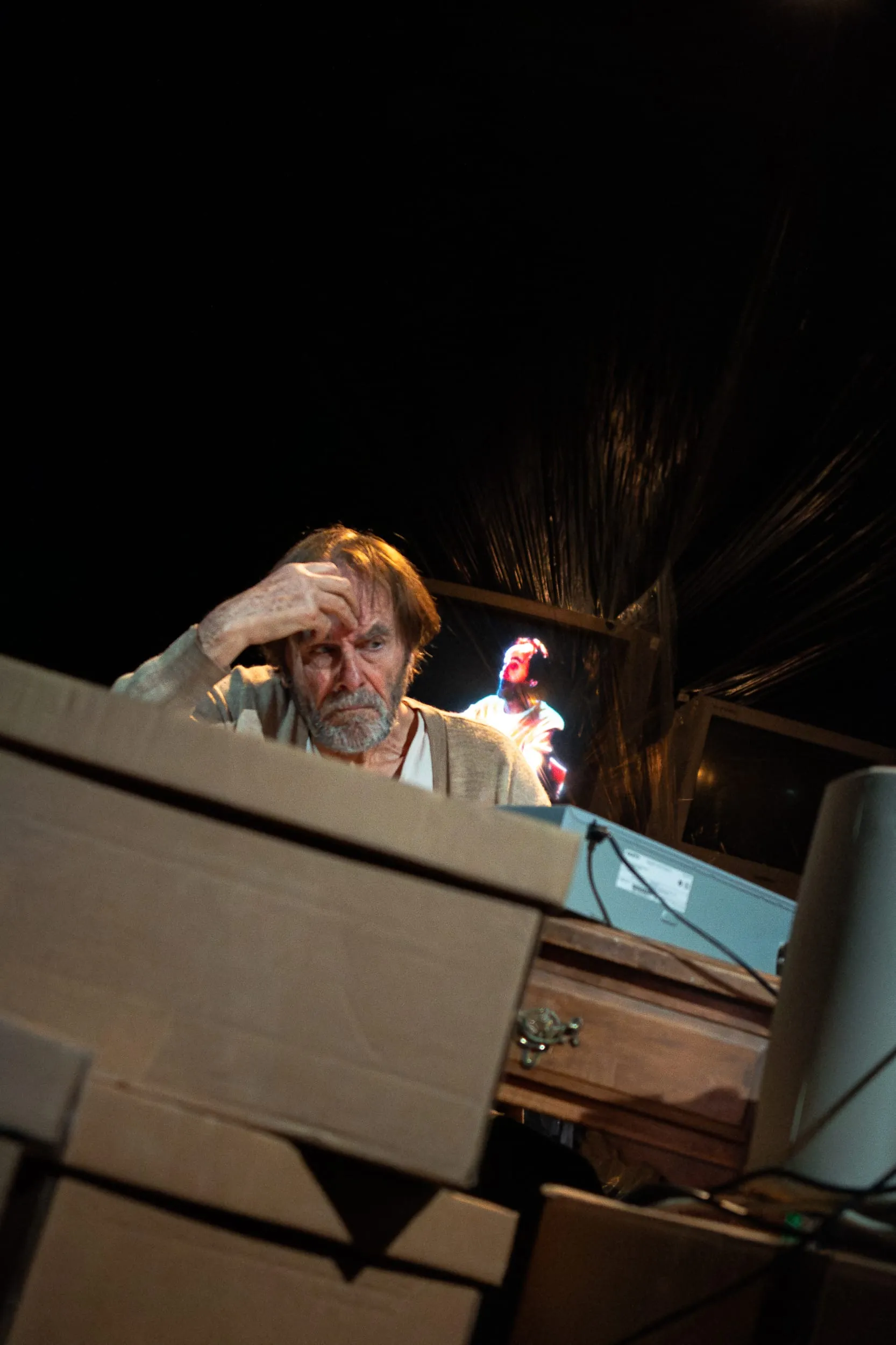 Krapp bows his head over the boxes while a red-lit face looms on a screen in the dark