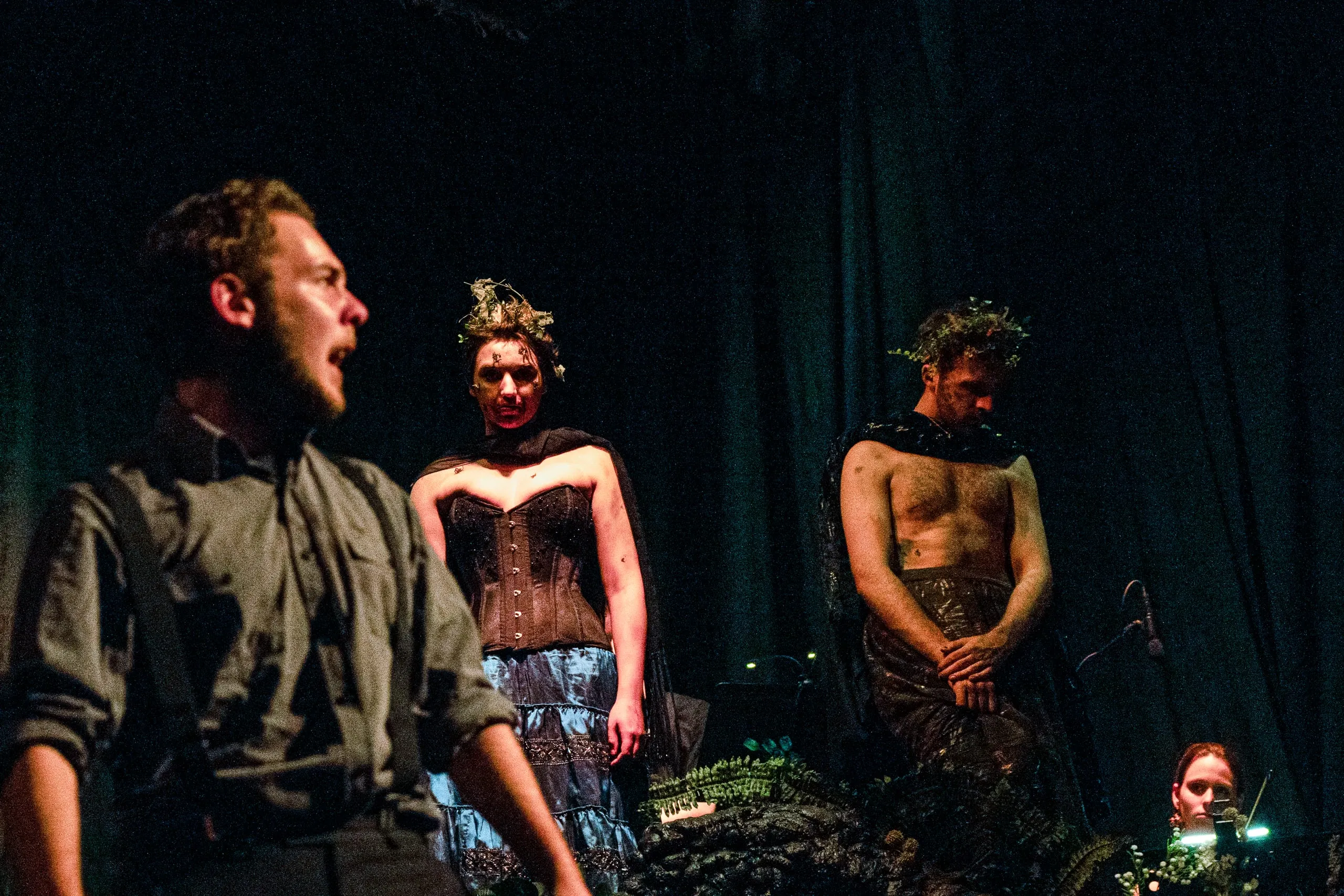 The witches — twig-crowned, corseted, draped in black — stand among the ferns as a soldier cries out in the foreground
