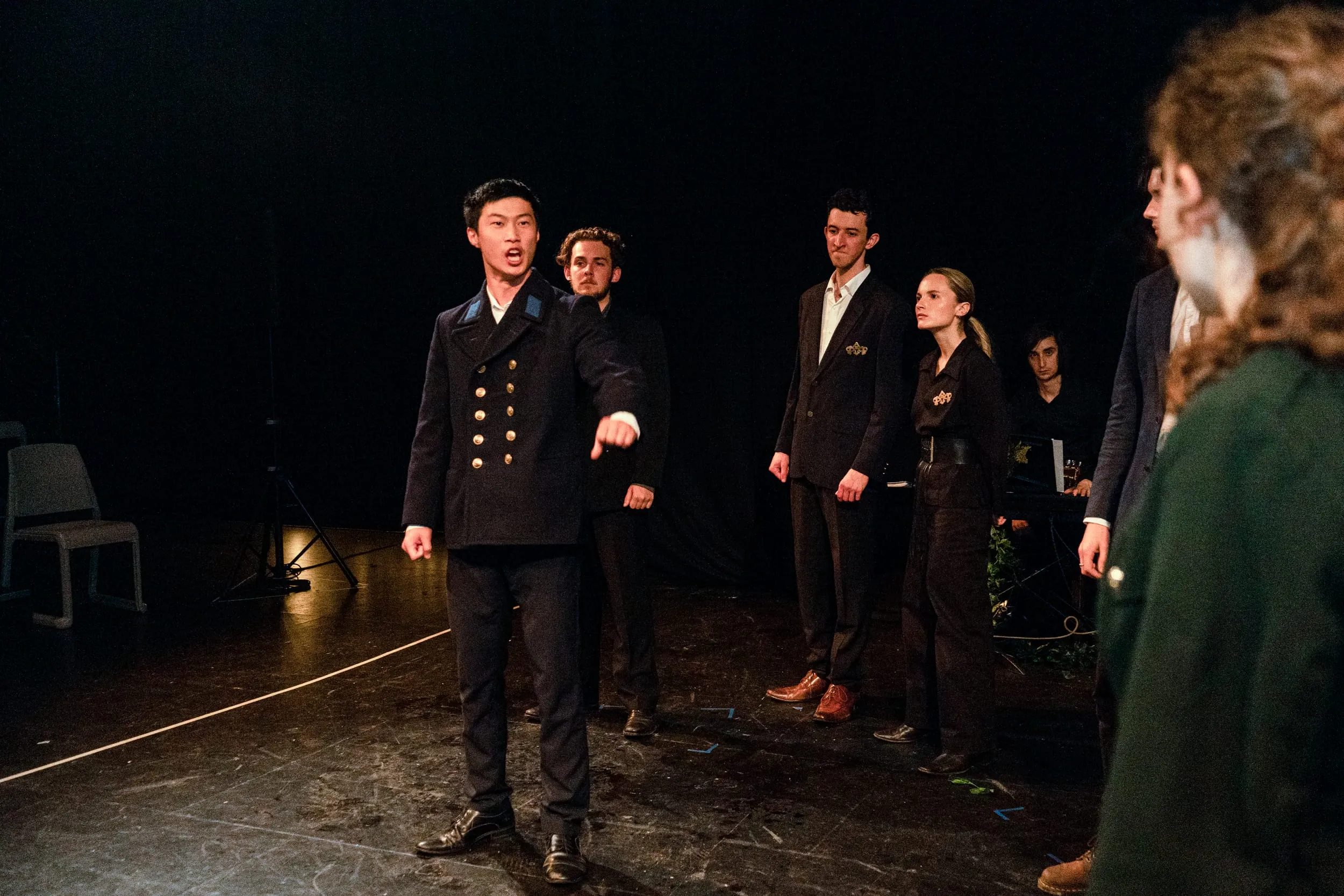Macbeth in a naval coat points accusingly downstage while the court watches in a line behind him