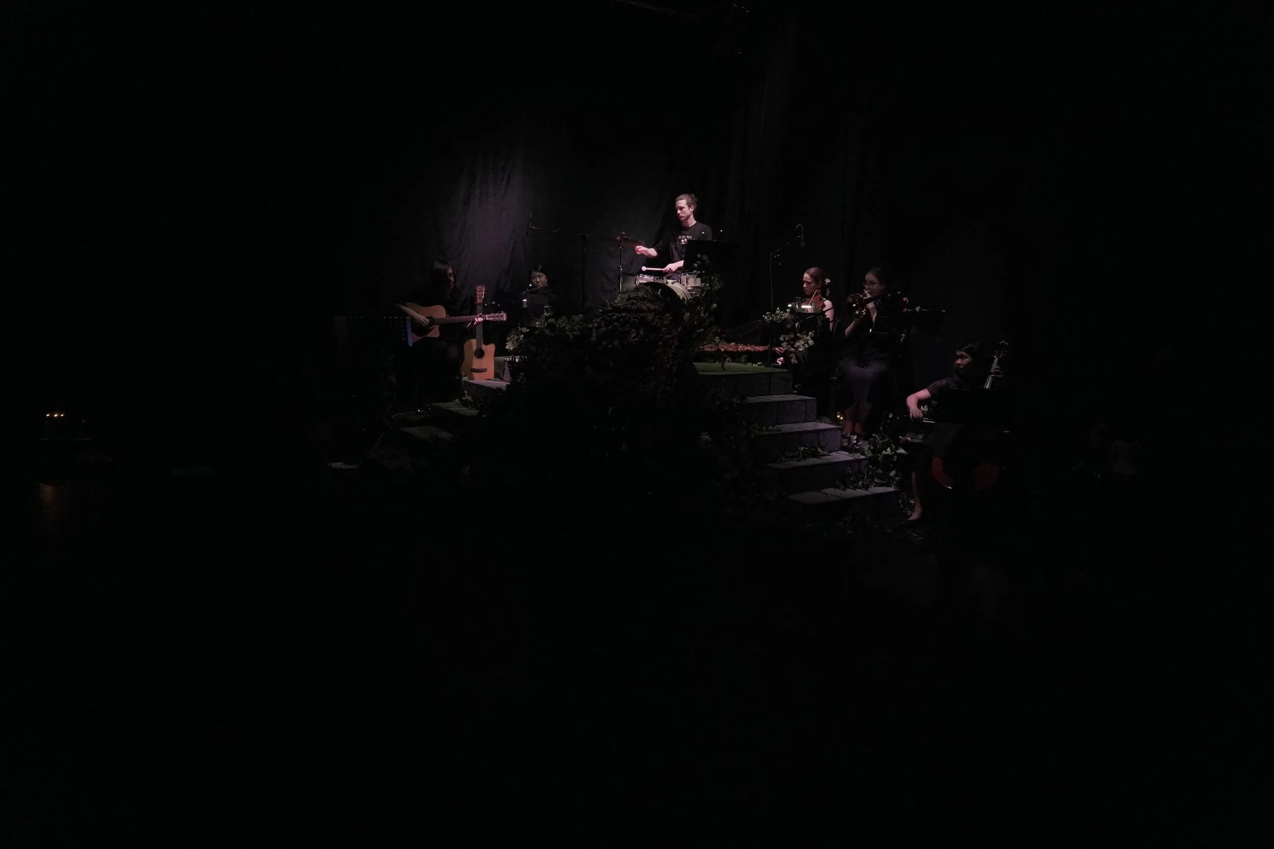 The full stage in near-darkness: a lone figure lit high on the steps, the band scattered in the gloom