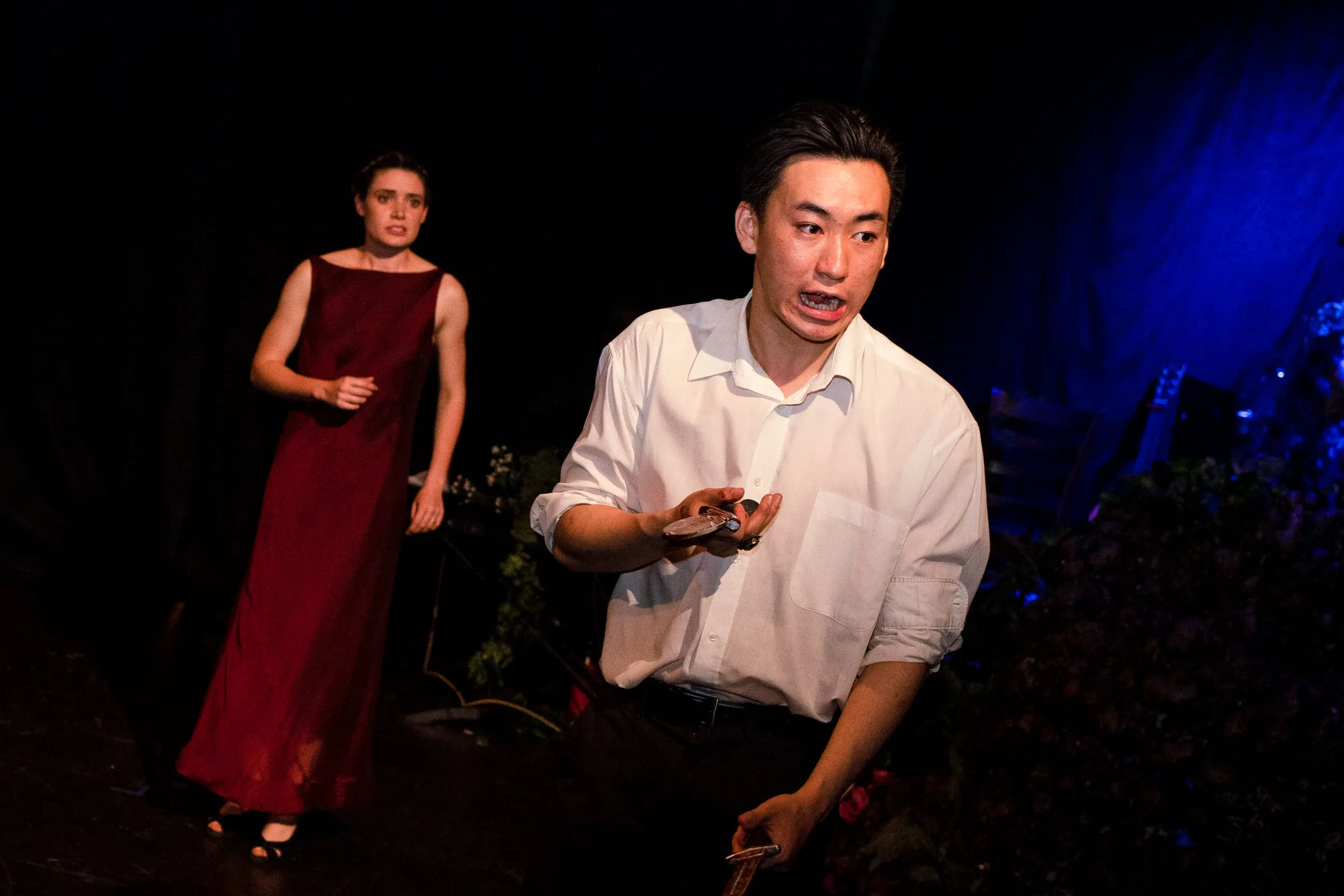Macbeth kneels among the foliage clutching the daggers, Lady Macbeth watching from the shadows in a deep-red gown