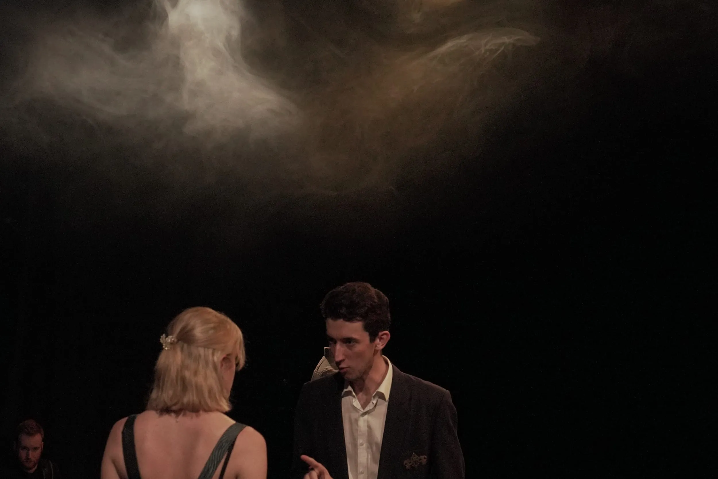 Two figures confer beneath a swirl of smoke caught in the stage light