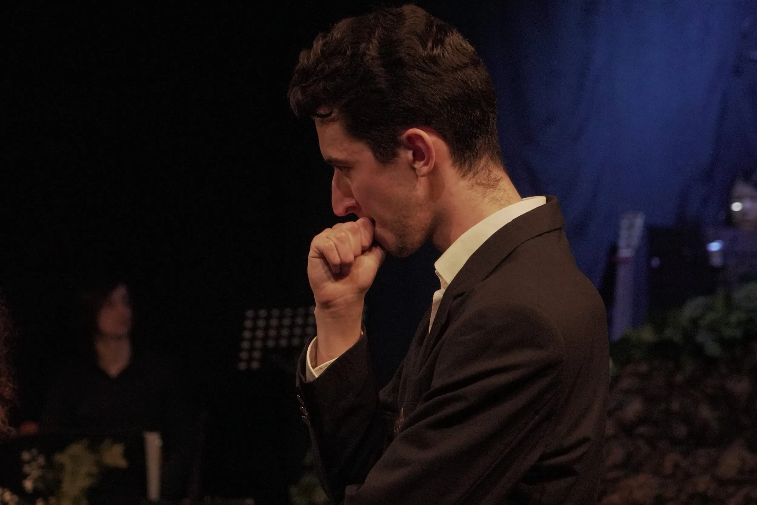 A suited figure presses a fist to his lips in profile, the band faint in the blue light behind