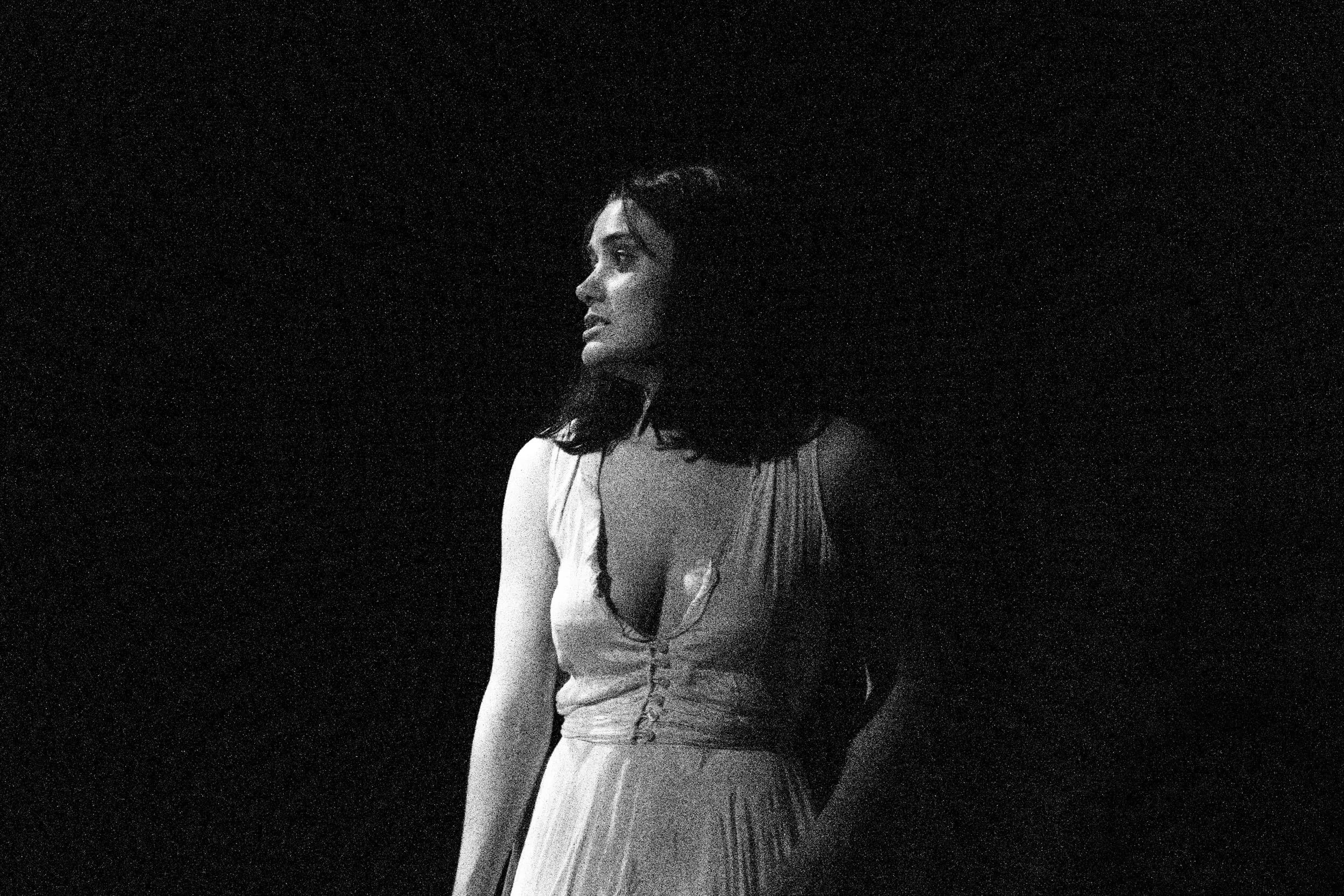 Black-and-white: Lady Macbeth stands alone in a pale gown, caught in a soft pool of light