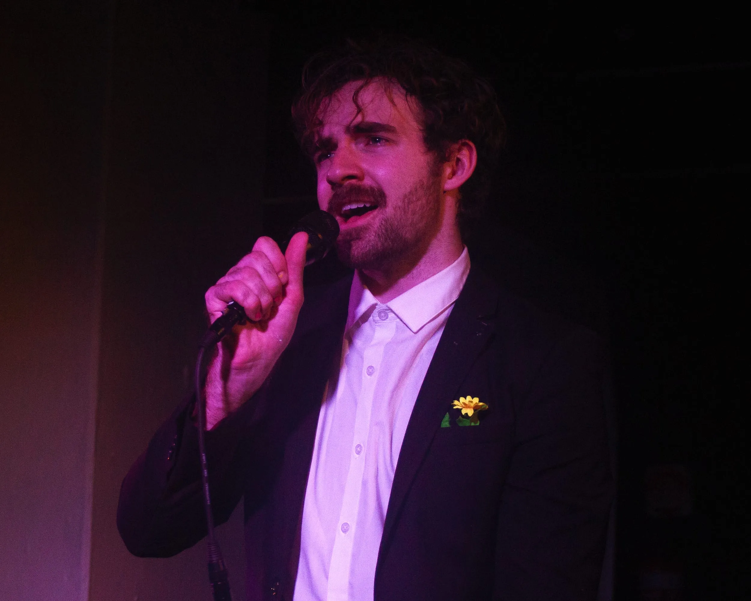 Close-up: the performer sings into the mic, a yellow daffodil pinned to his jacket