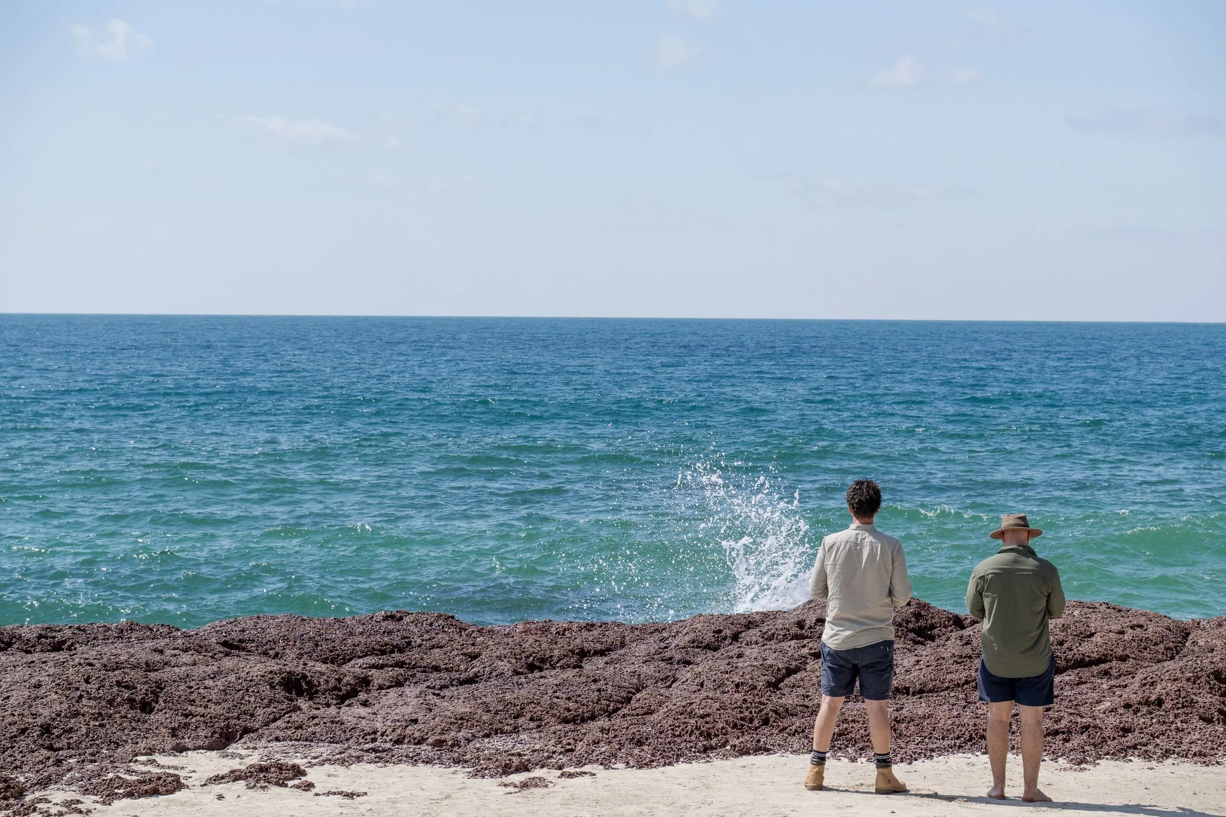 Two figures watch spray burst on the rocks at the water’s edge