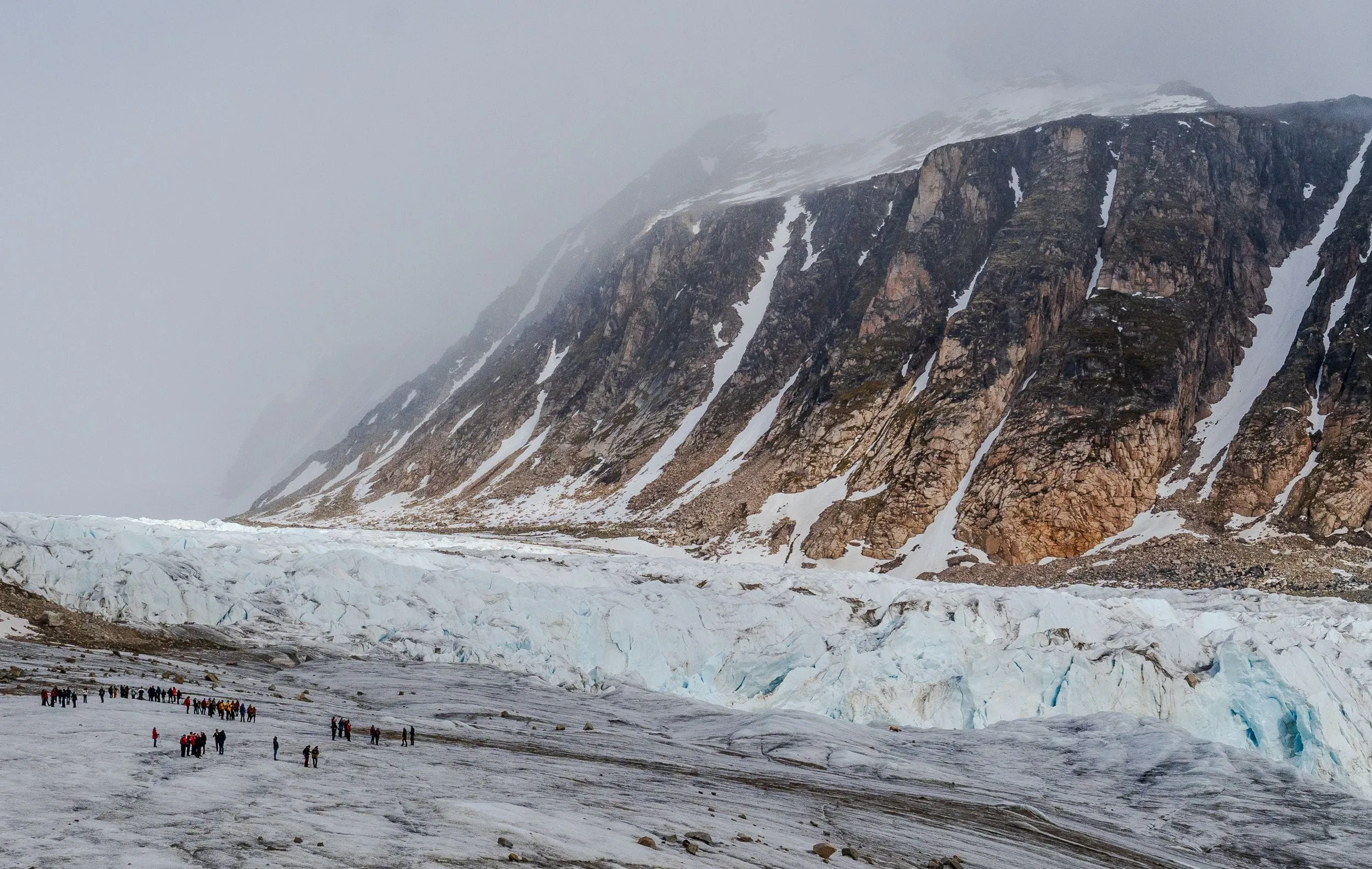 Walkers in single file dwarfed by a glacier front and dark cliffs, Svalbard