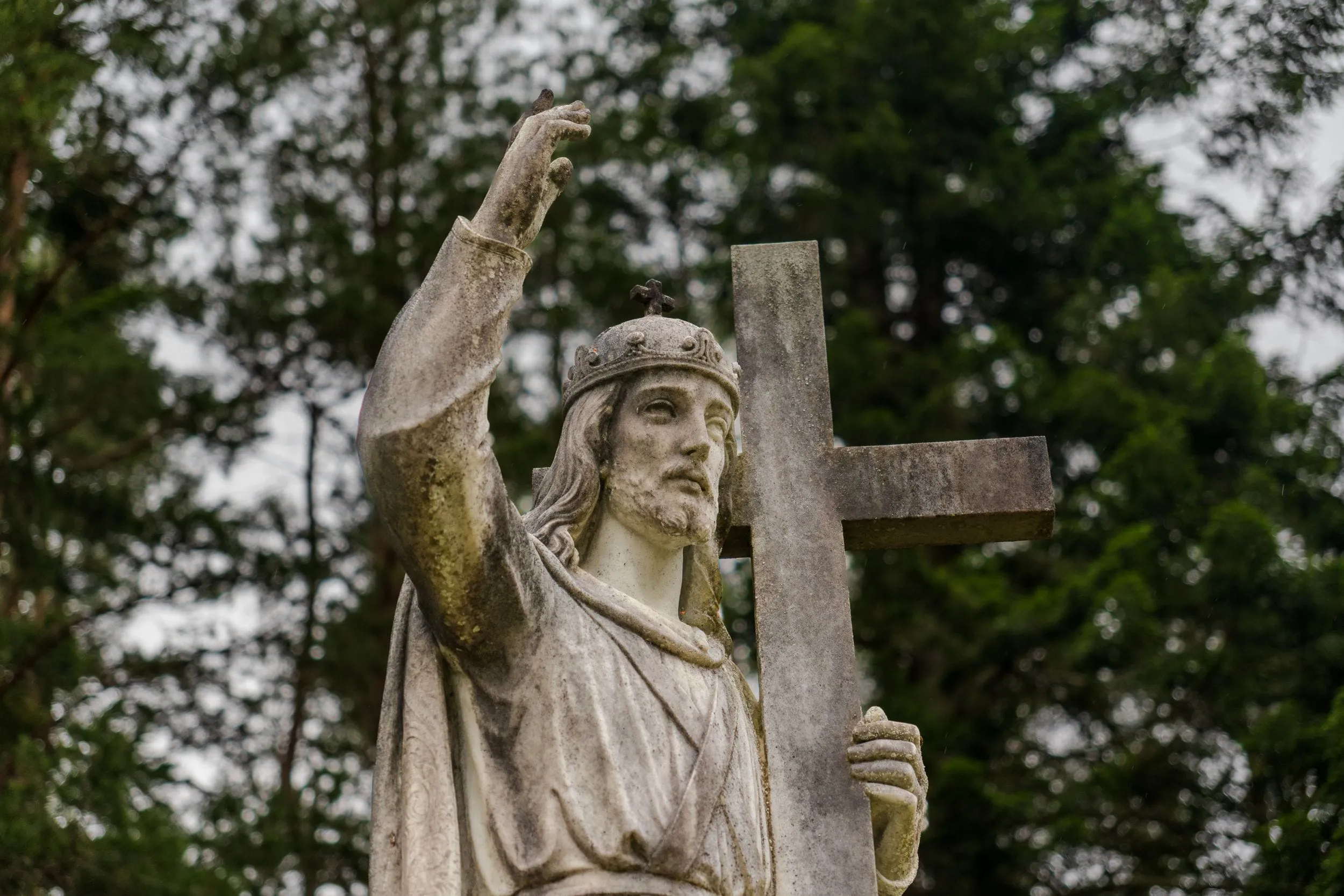 A stone figure raising a hand beside its cross, framed by dark pines