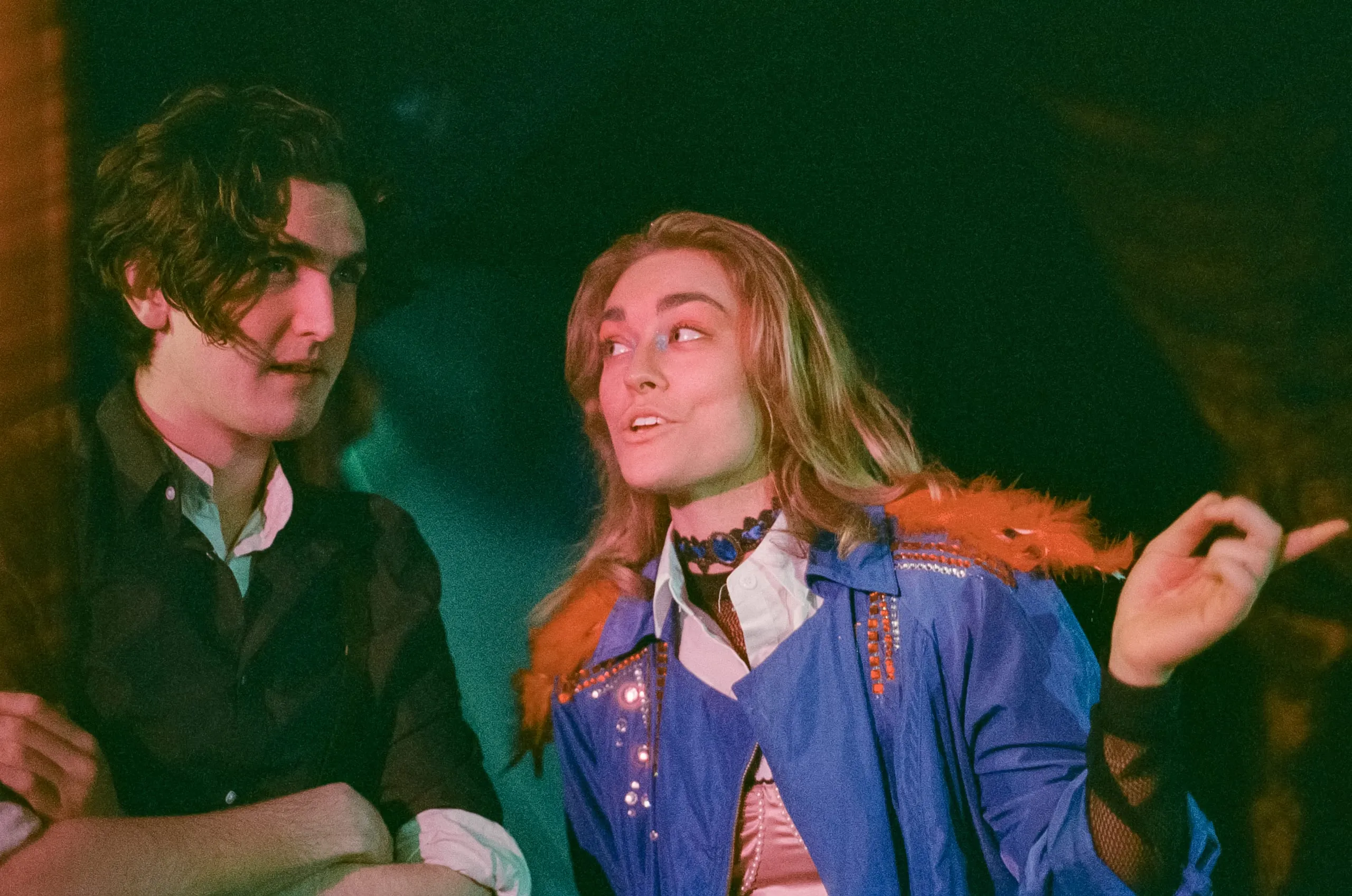 The Player, in a jewelled blue jacket and feather boa, shares a look with a wary companion under green light