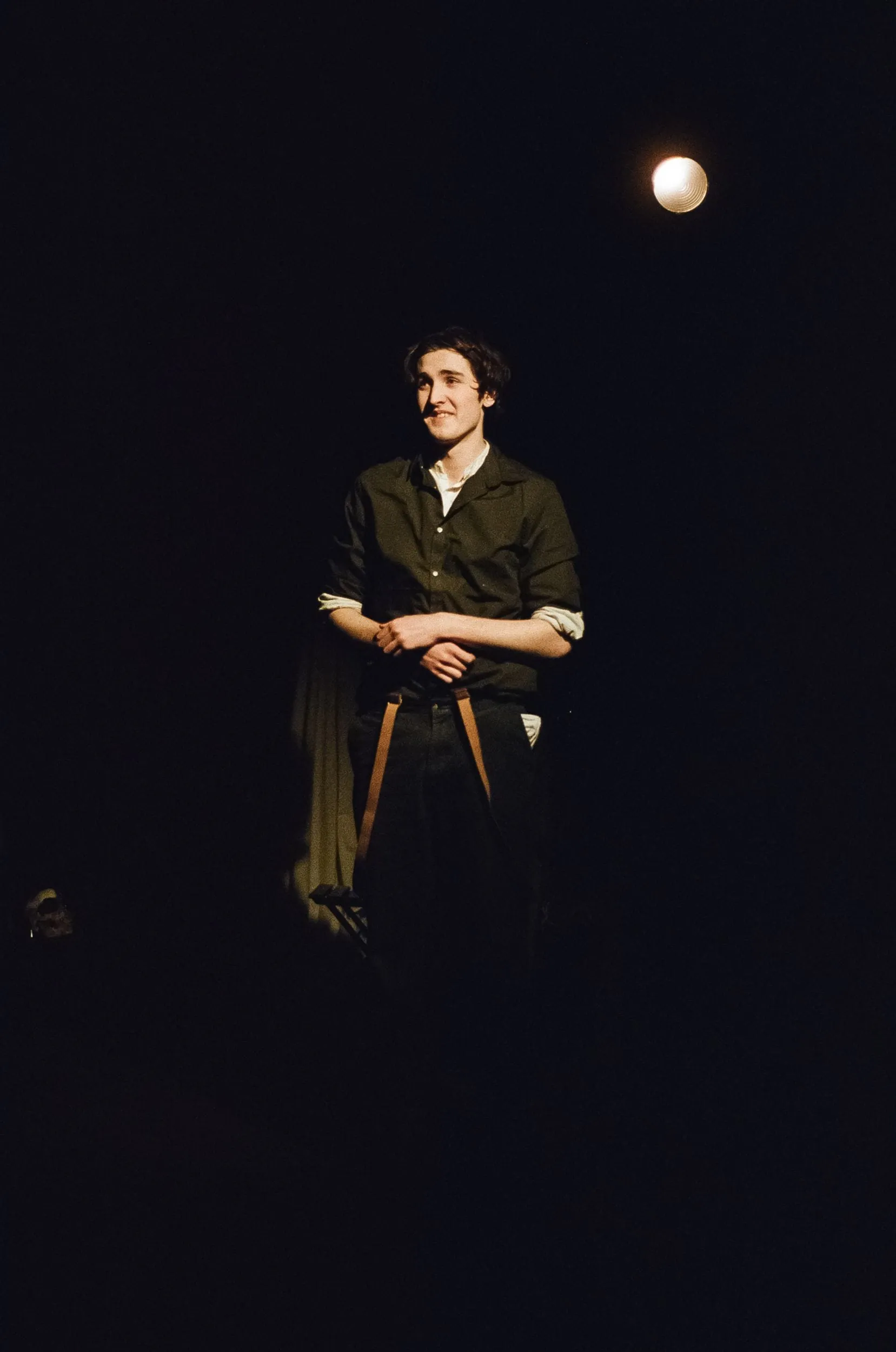 A performer smiles in a small island of light, hands folded at his waist