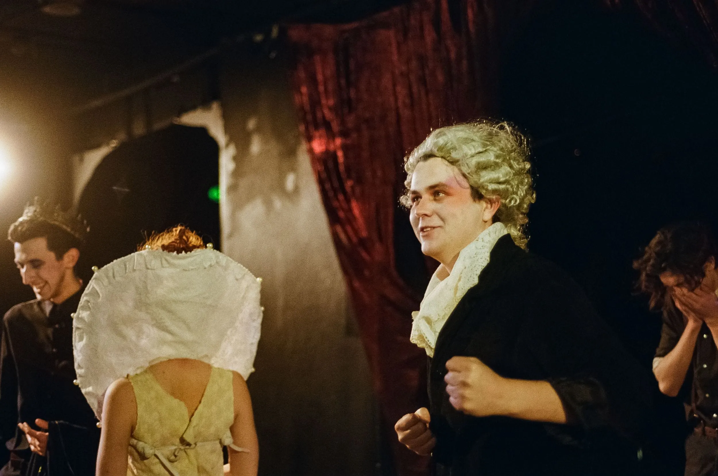 A white-wigged player grins amid the troupe before a red curtain