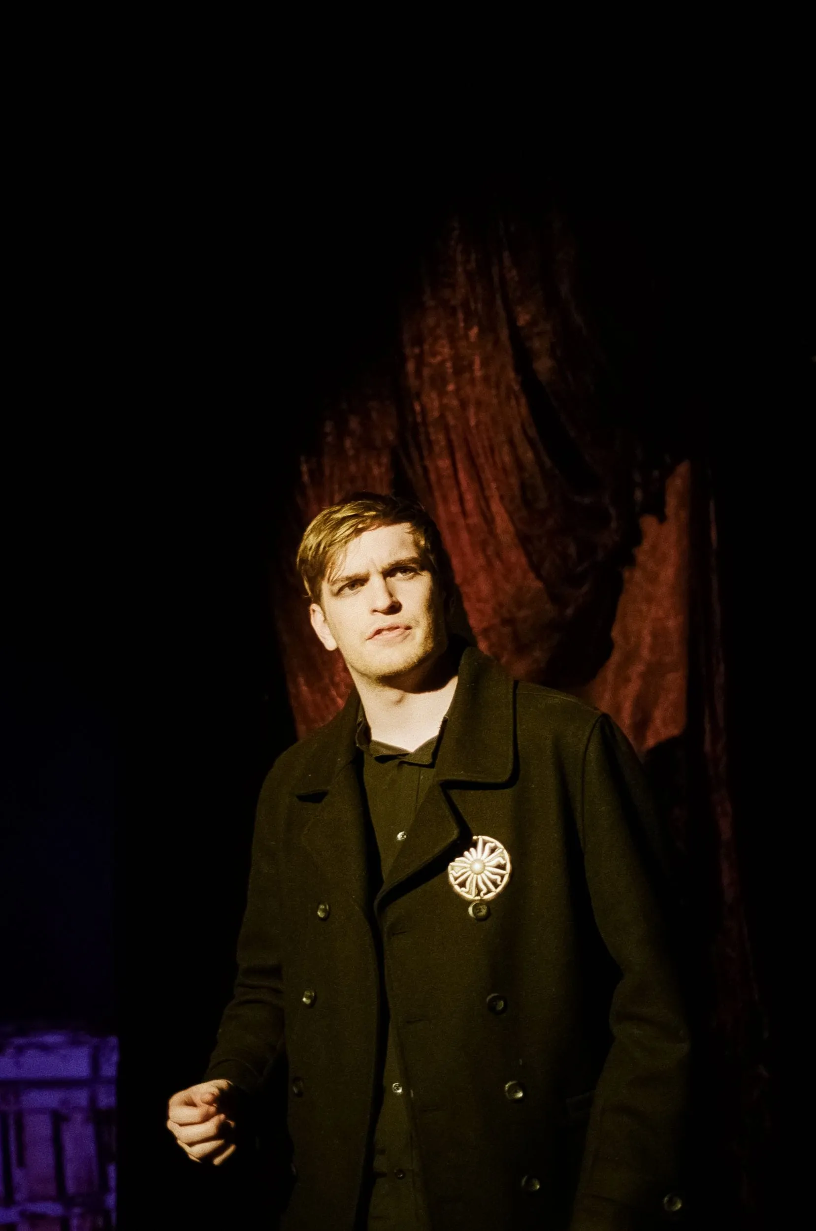 A coated figure wearing a medallion stands wary before the red curtain