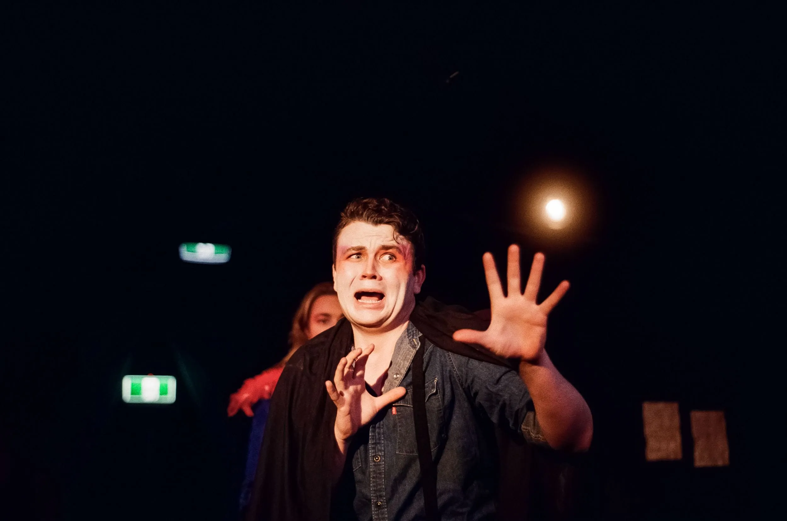 A cloaked performer recoils white-faced, hand splayed against the light