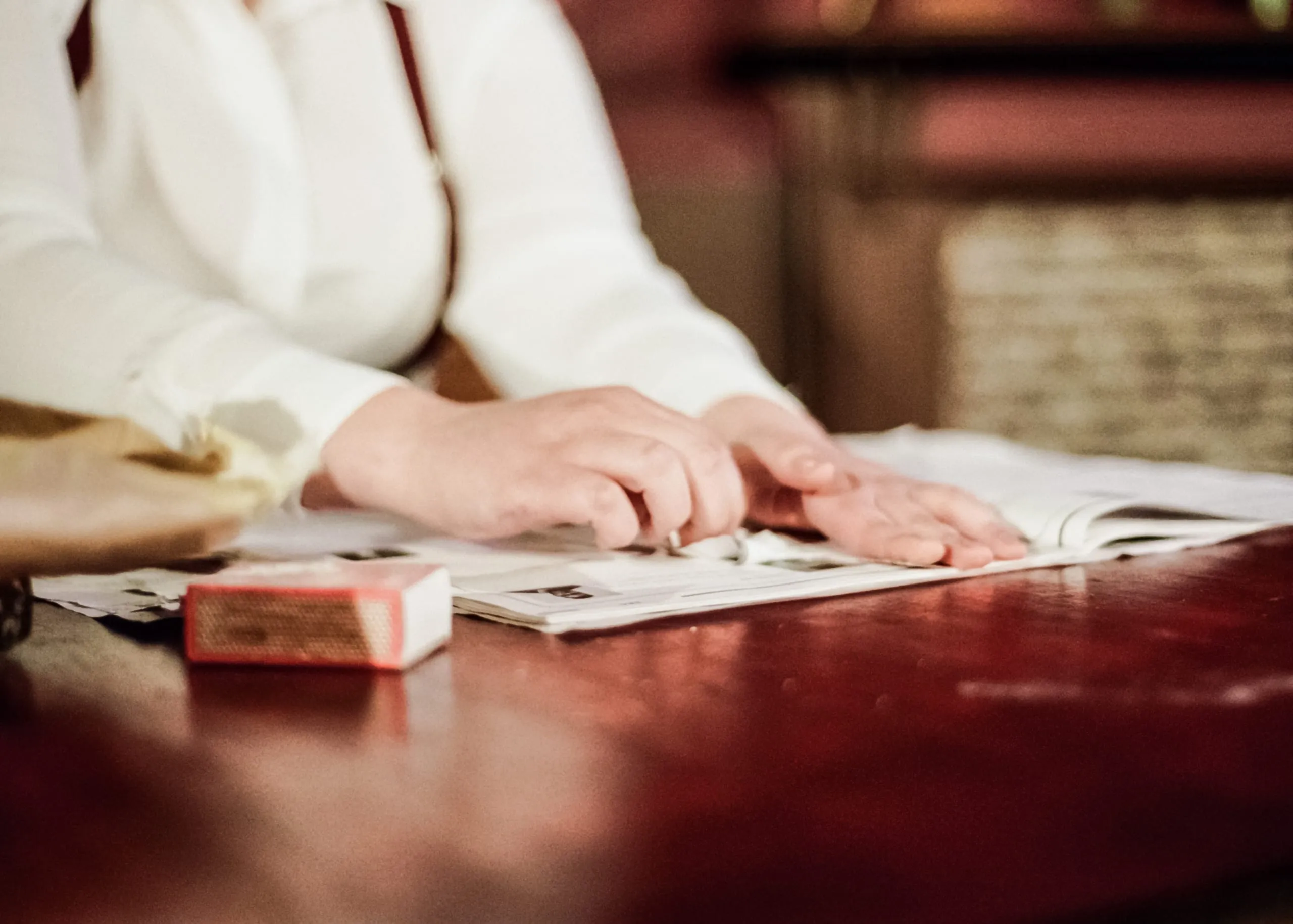 Close-up: hands smooth a newspaper open on the deep-red table beside a box of matches