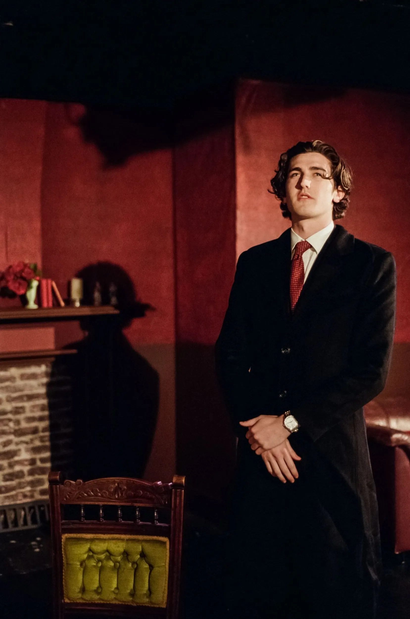 Goldberg stands composed in the red-walled parlour beside a green-cushioned chair, hands clasped at his watch